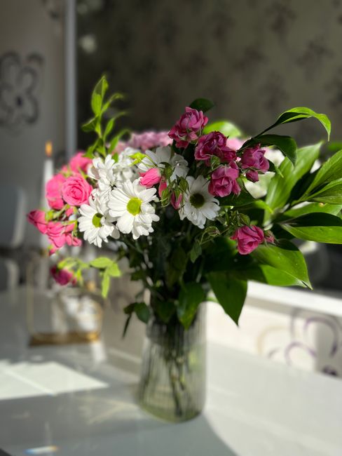 A fresh floral bouquet arranged in a clear glass vase sits on a white surface indoors, featuring a mix of pink roses, white daisies with yellow centers, and pink bougainvillea flowers with vibrant gre