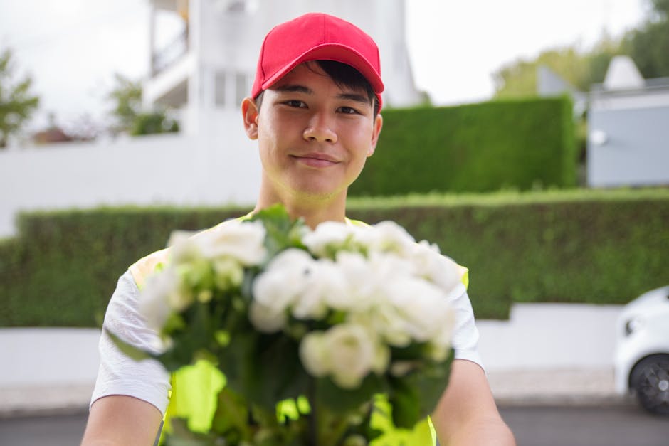 A delivery person wearing a red cap and a high-visibility vest is holding a bouquet of fresh white flowers with green foliage, extending it towards the camera. The bouquet appears to include roses and