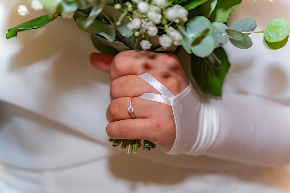 A close-up of a bride's hand holding a bridal bouquet consisting of white and green flowers, including baby's breath and eucalyptus leaves, wrapped in white satin ribbon. The bride is wearing a weddin