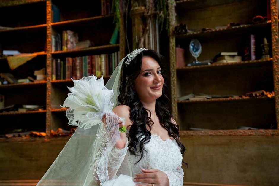 A bride with long, dark, wavy hair framed by a floral headpiece and veil, holding a bouquet of white lilies wrapped in sheer white tissue and lace, wearing a white lace wedding gown with off-the-shoul
