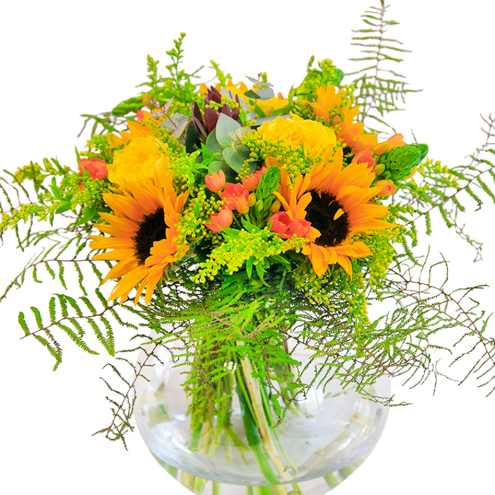 Encountering this bouquet is like stepping into the height of summer in Ardleigh Green, where gardens overflow with colour and laughter. Dominating the scene are majestic sunflowers-their round, golden faces and chocolate centers reminiscent of joyful afternoons under wide Essex skies. Nestled close, classic yellow garden roses spiral open in generous layers, recalling horticultural tradition and the area's love for elegant blooms. Scarlet Hypericum berries, glossy and round, peek out among cascades of forest-green ferns and airy filler, mimicking the playful vibrancy of Ardleigh Green's high street on market day. The round fishbowl vase, perfectly clear and modern, cradles every stem, while the water inside glimmers in sunlight like reflections on the River Ingrebourne. Textural interplay unfolds as feathery greens soften bold blooms, and silver-green eucalyptus leaves inject a fresh, modern twist. A faint, comforting floral scent lingers, summoning images of parks, picnics, and garden walks from Ardleigh Green Drive to local sanctuaries. Presented as a cheerful centerpiece for celebrations, thoughtful congratulations, or simply a way to fill a room with joy, this arrangement captures the essence of Ardleigh Green's warm community and its florists' skilled hands.
